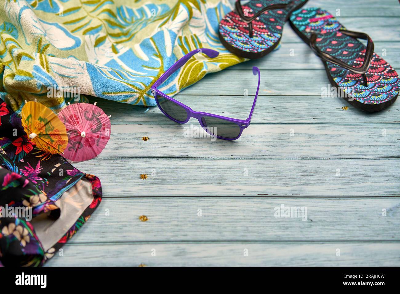 Lila Sonnenbrille mit zwei kleinen orange-rosa Sonnenschirmen, ein Handtuch mit tropischem Muster und Flip-Flops mit tropischem Muster auf einem Holztisch. Stockfoto