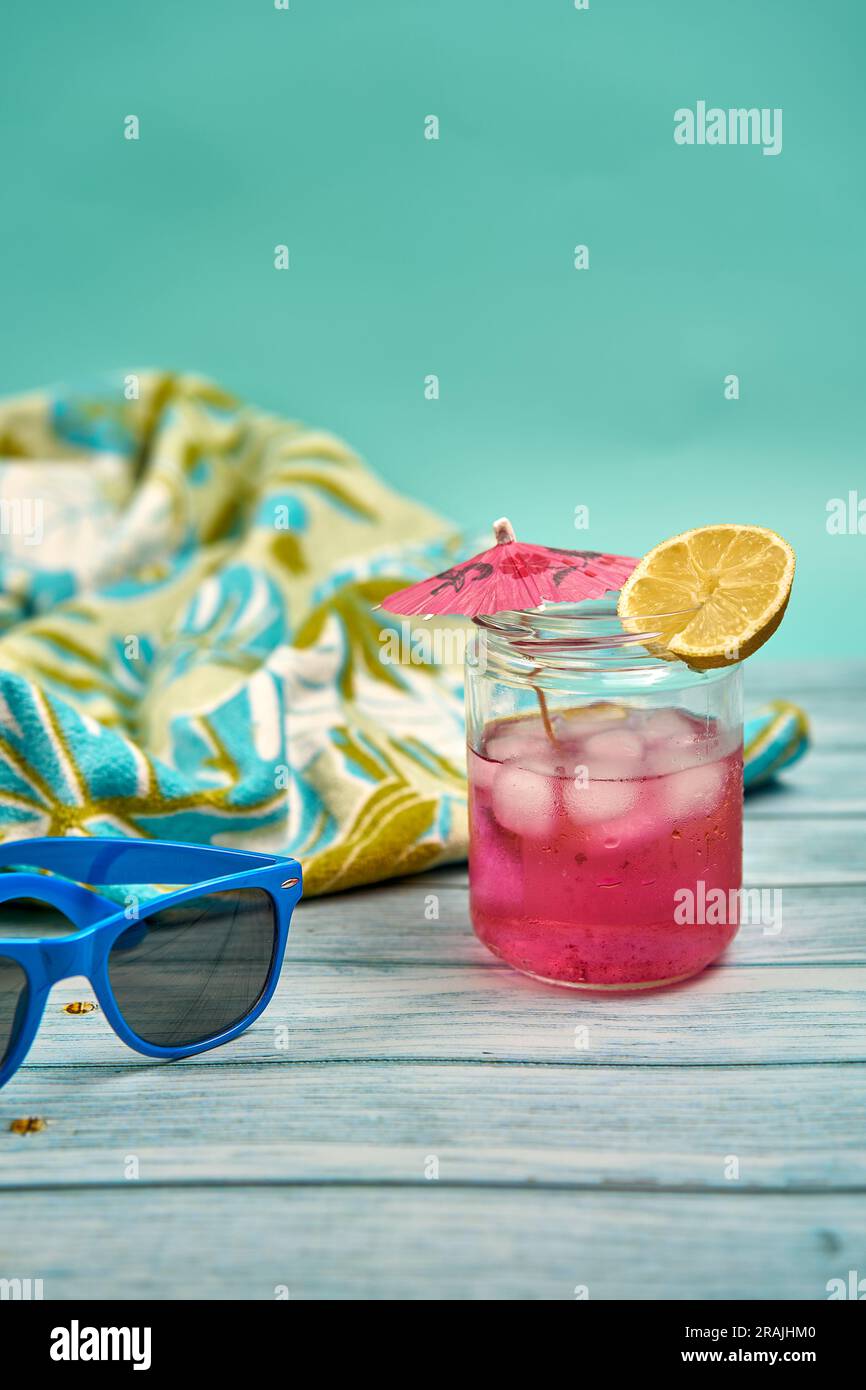 Erfrischendes Sommergetränk mit viel Eis, Schirm und ein Stück Zitrone, blaue Sonnenbrille und tropisch gemustertes Strandtuch auf einem Holztisch und einem Stockfoto