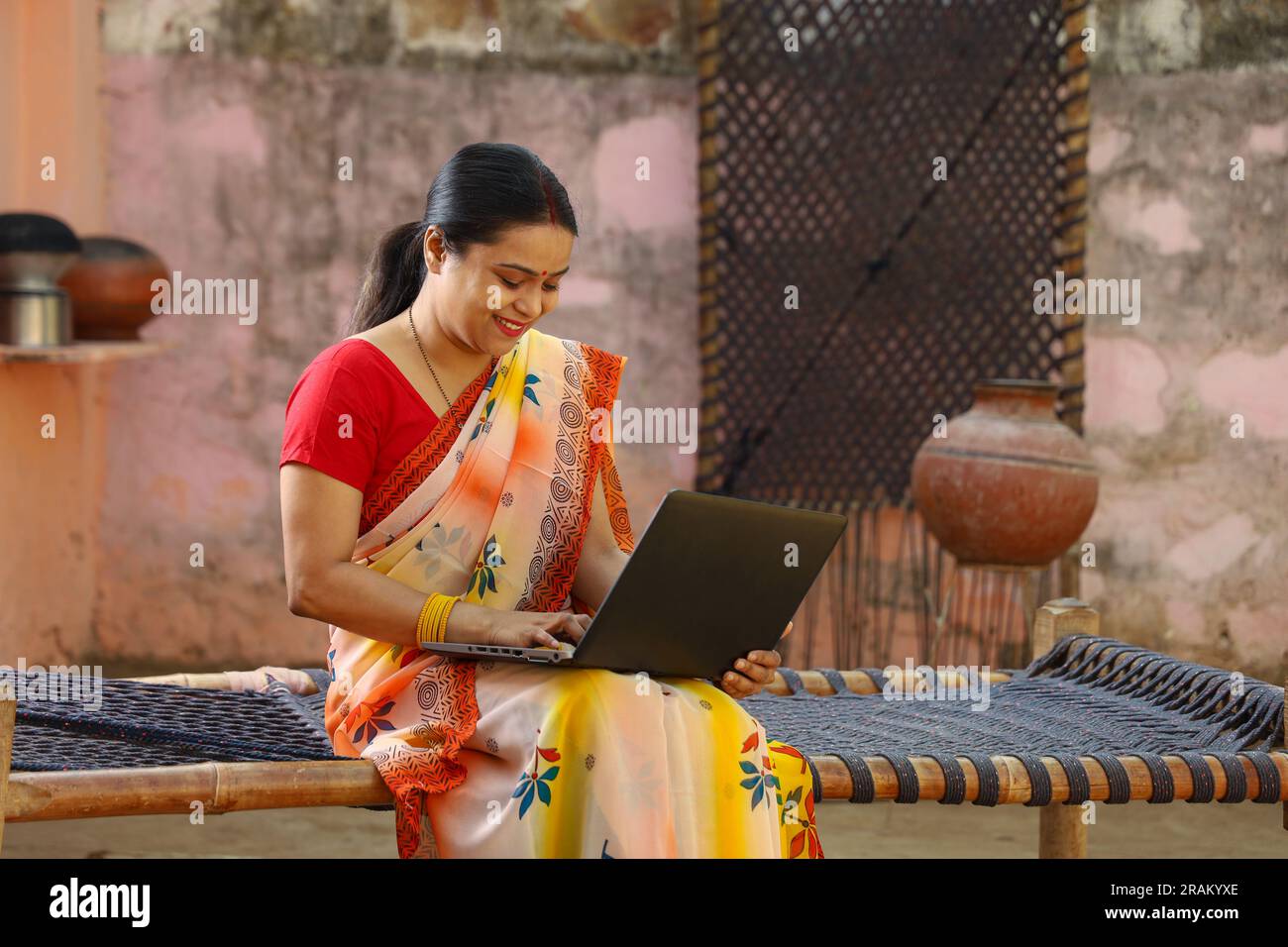 Eine glückliche Frau aus dem indischen Dorf in Saree, die den Laptop benutzt, der vor dem Haus sitzt und Daumen nach oben macht. Eine ländliche Indianerin, die auf einem Kinderbett sitzt. Technologiekonzept. Stockfoto