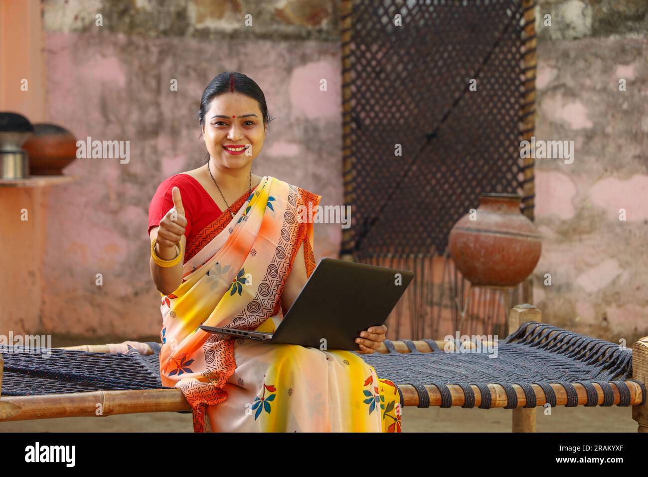 Eine glückliche Frau aus dem indischen Dorf in Saree, die den Laptop benutzt, der vor dem Haus sitzt und Daumen nach oben macht. Eine ländliche Indianerin, die auf einem Kinderbett sitzt. Technologiekonzept. Stockfoto