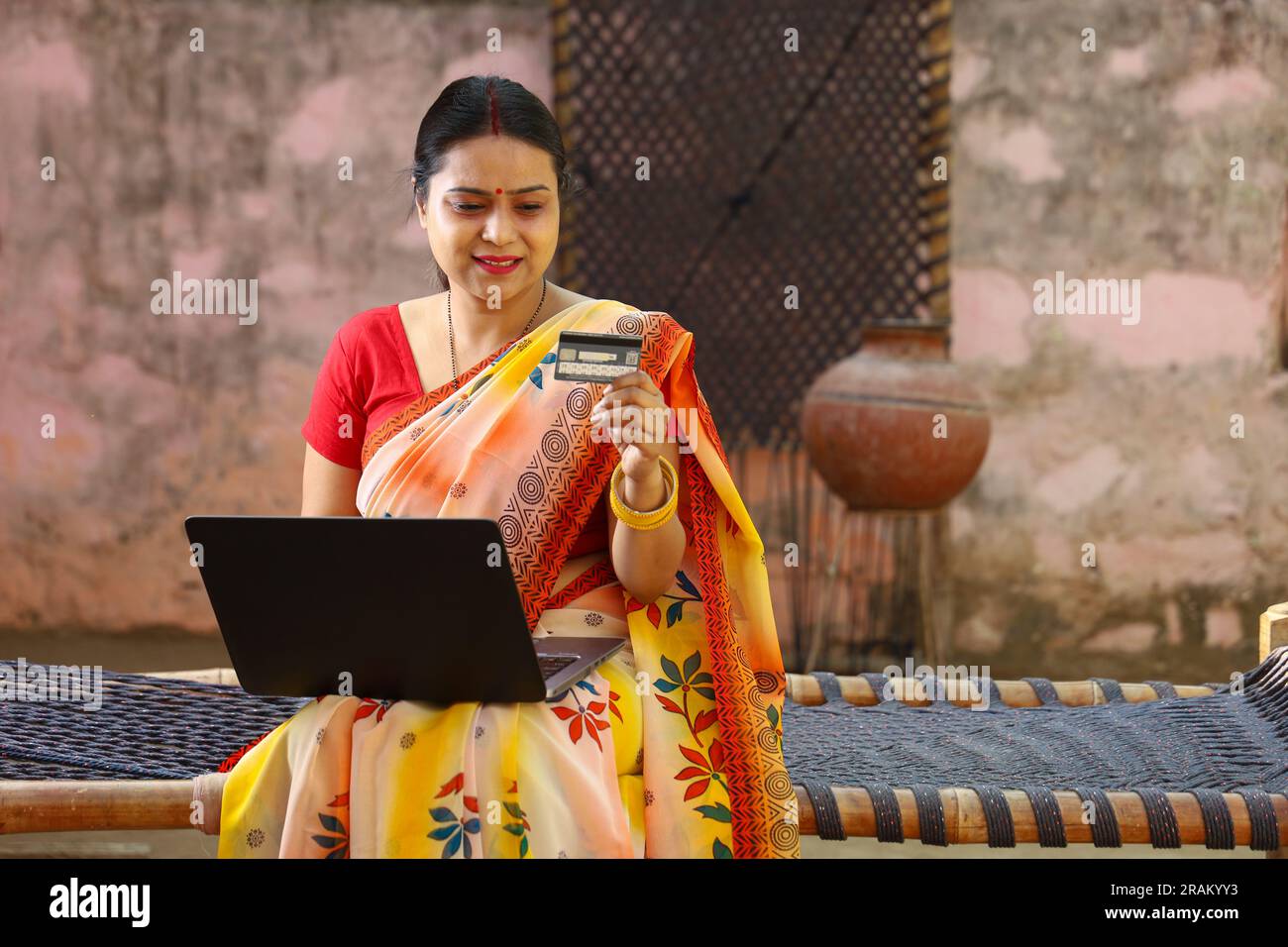 Glückliche Frau aus dem indischen Dorf in Saree, die den Laptop vor dem Haus benutzt und online mit Kreditkarte bezahlt hat. Stockfoto