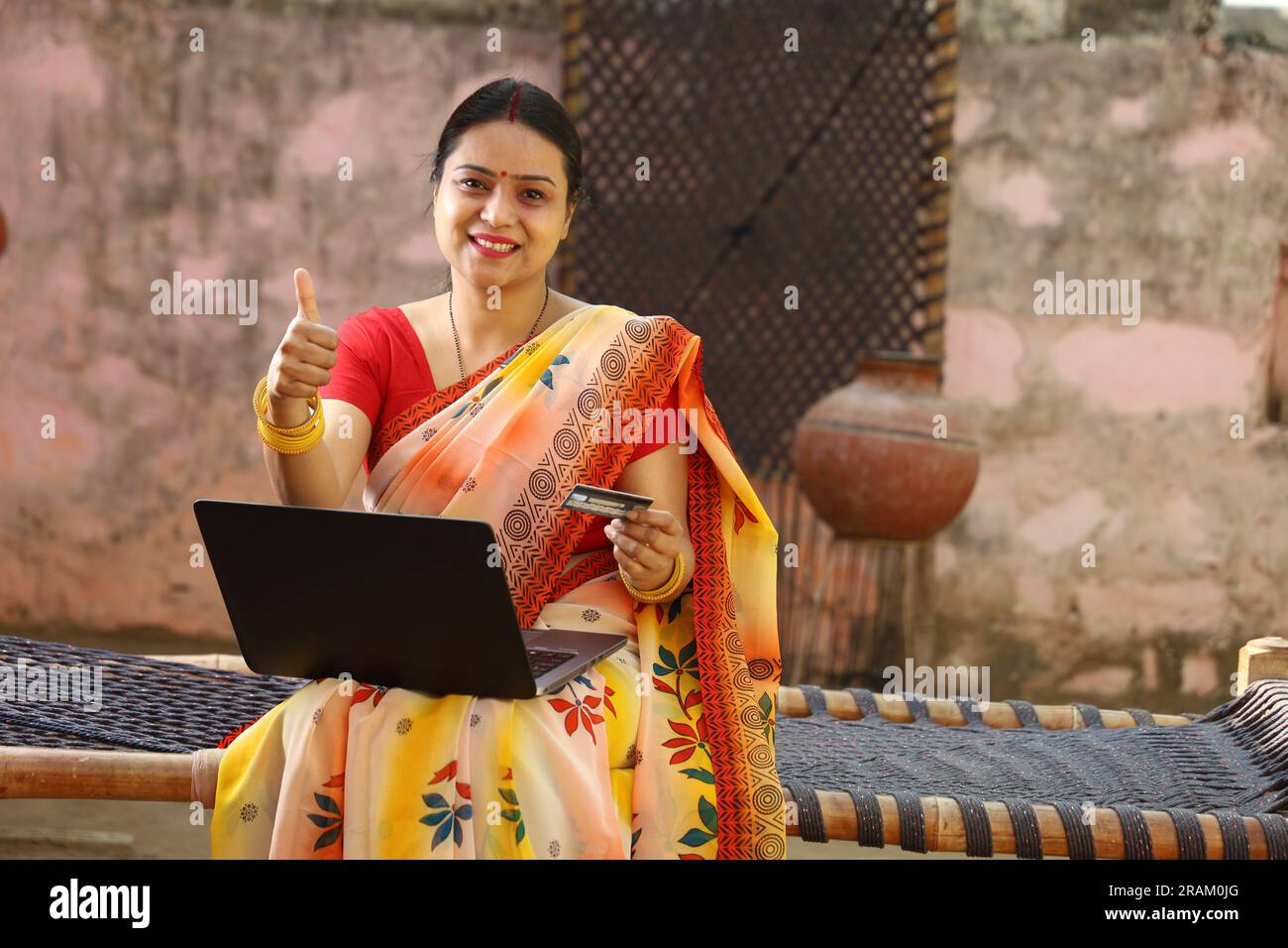 Glückliche Frau aus dem indischen Dorf in Saree, die den Laptop vor dem Haus benutzt und online mit Kreditkarte bezahlt hat. Stockfoto
