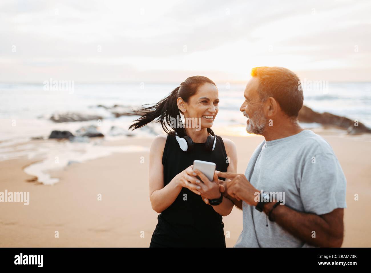 Die fröhliche ältere weiße Dame und der Mann in Sportbekleidung nutzen die Telefon-App, genießen Training und aktiven Lebensstil am Strand Stockfoto