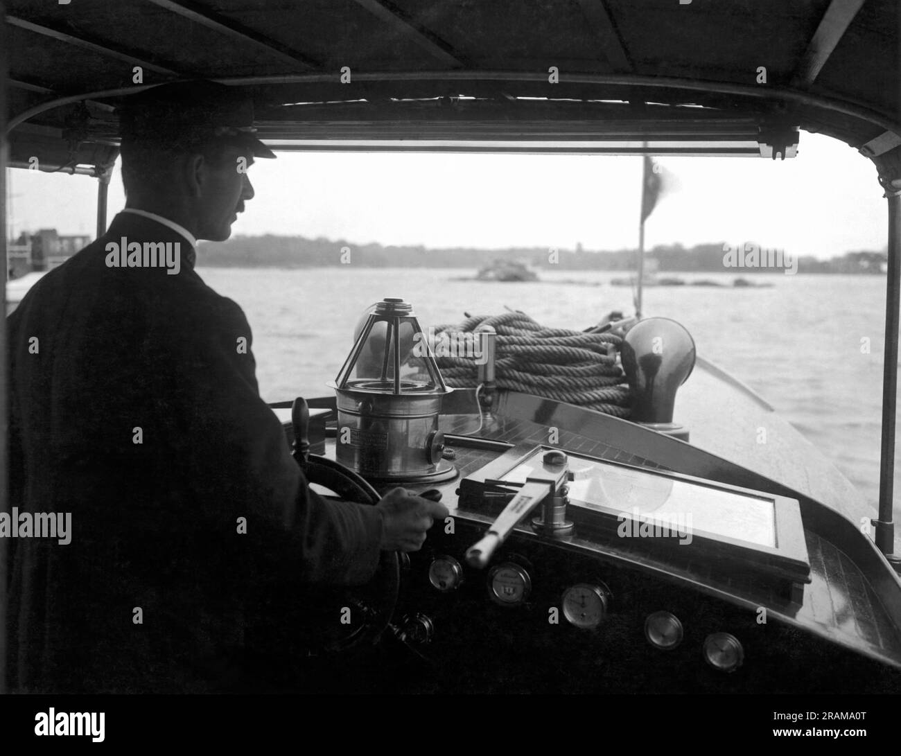 Vereinigte Staaten: c. 1925. Ein Mann in einem überdachten Cockpit führt sein großes Motorboot auf einen Wasserweg Stockfoto