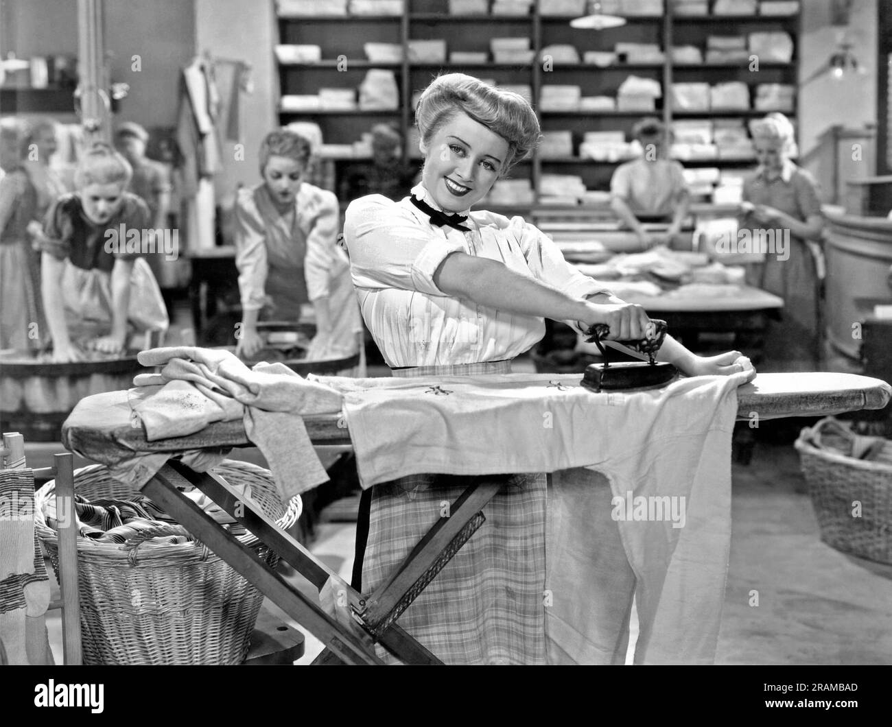 Vereinigte Staaten: c. 1930. Eine lächelnde Frau bügelt ihre Kleidung fröhlich mit einem flachen Bügeleisen in der Wäsche. Stockfoto
