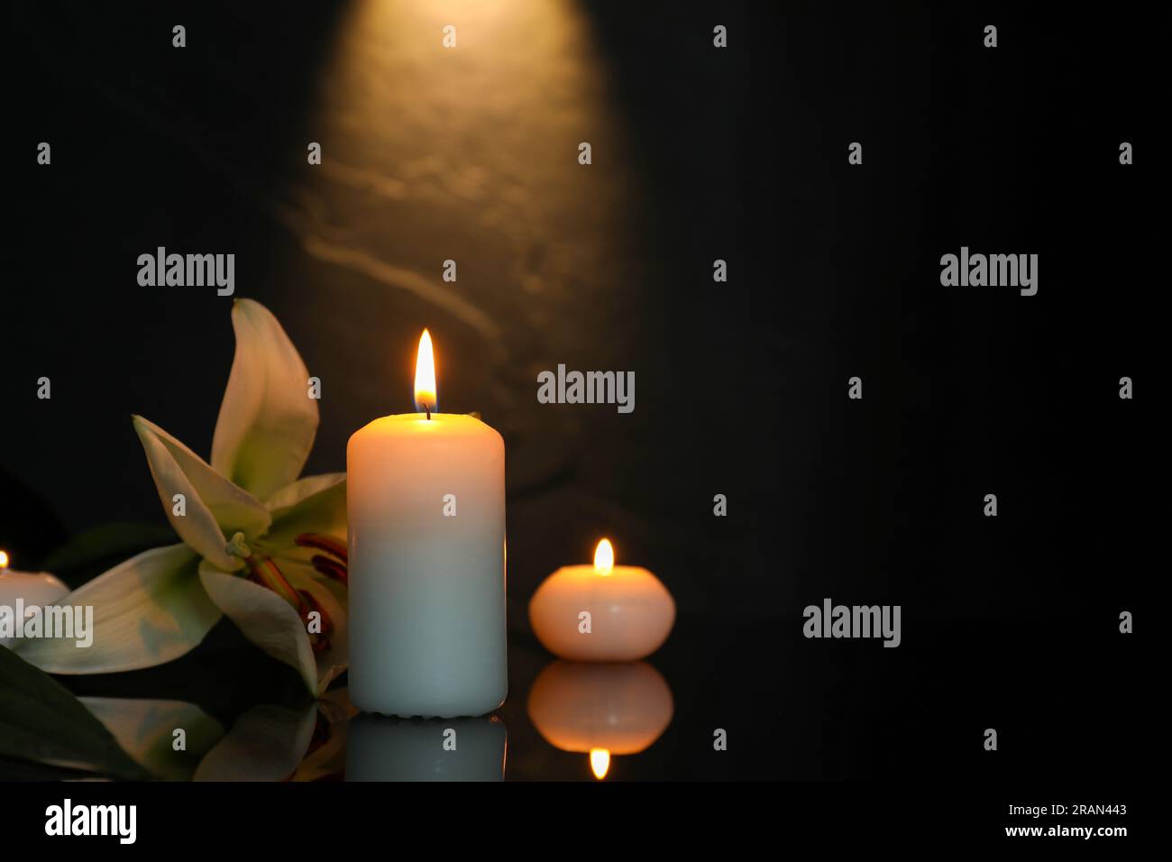 Weiße Lilie und brennende Kerzen auf schwarzer Spiegeloberfläche bei Dunkelheit, Platz für Text. Begräbnissymbole Stockfoto