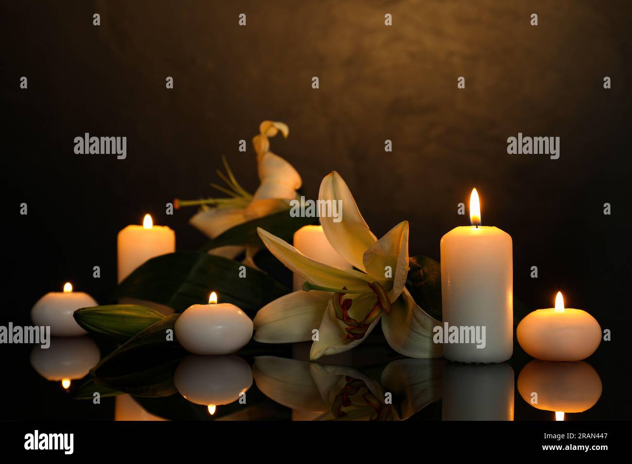 Weiße Lilien und brennende Kerzen auf schwarzer Spiegeloberfläche bei Dunkelheit, Platz für Text. Begräbnissymbole Stockfoto