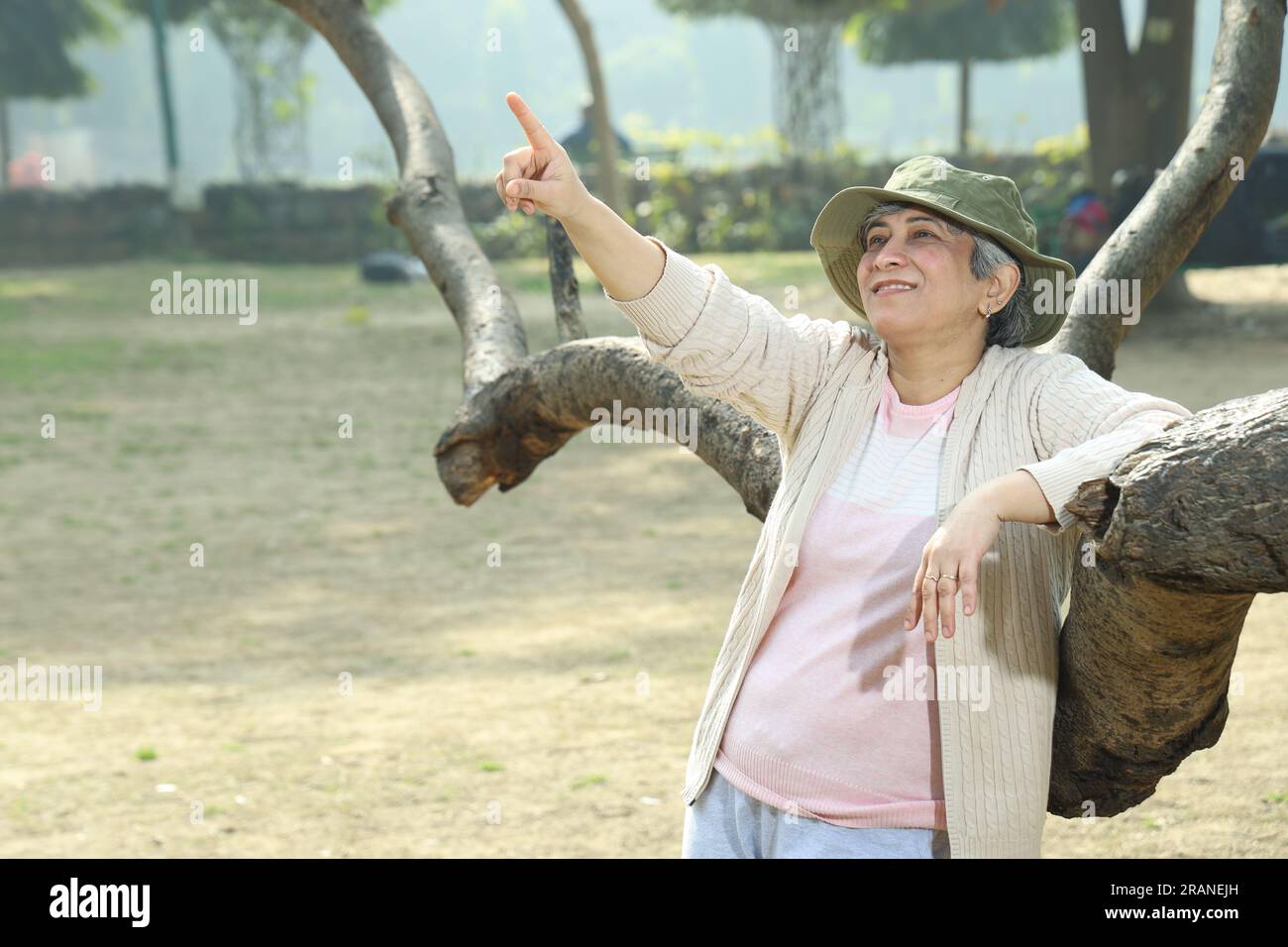 Porträt einer gesunden indischen fröhlichen Frau mittleren Alters im Park, die neben dem Baum steht und Spaß hat. Sie trägt tagsüber einen Hut. Stockfoto