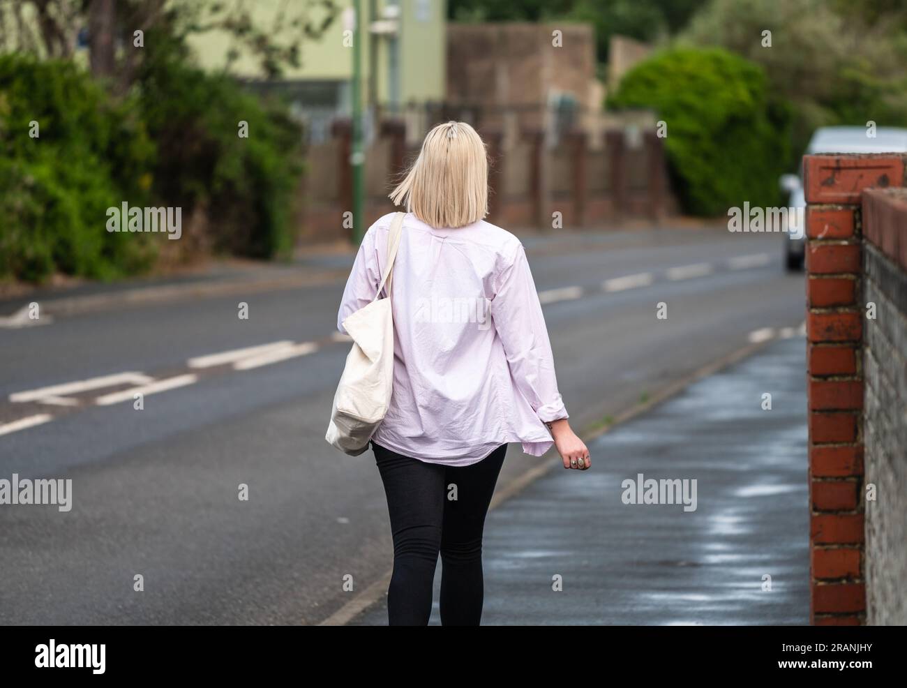 Das Heck einer jungen, stilvollen Dame mit blondem Haar, rosa Oberteil und Handtasche, die auf einem Bürgersteig in Großbritannien davonläuft. Stockfoto