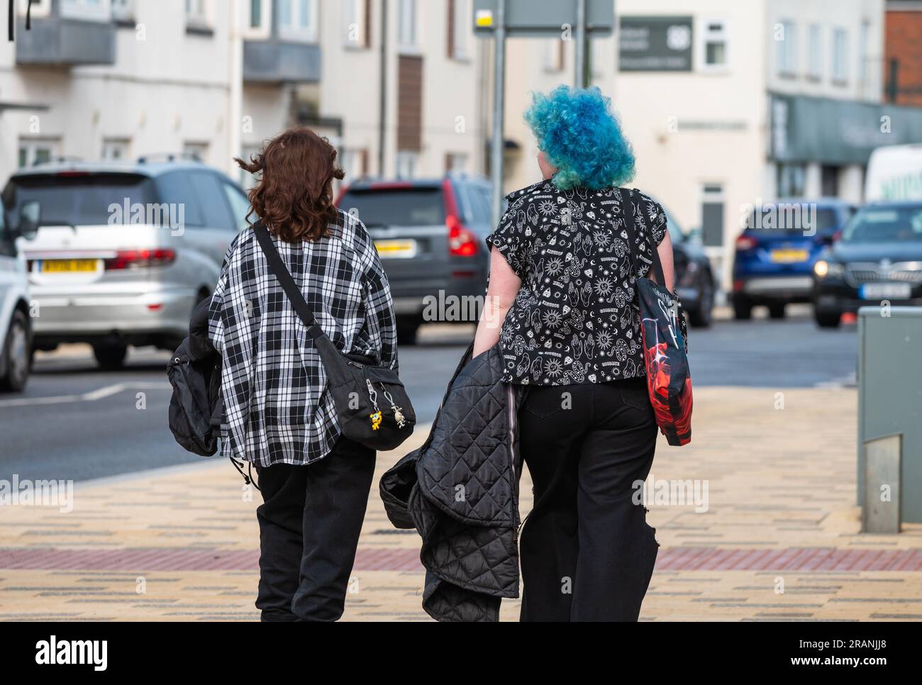 Zwei junge, moderne Frauen, möglicherweise Freunde, eine mit bunten blauen Haaren, die im Sommer in England auf einem Bürgersteig spaziert. Stockfoto