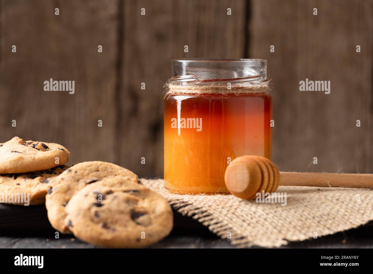 Honig in offenen Gläsern und in einer Schüssel, Dipper und hausgemachte Kekse mit Schokoladenstückchen auf Holzhintergrund. Hausgemachte Kuchen und Honig zum Frühstück. Stockfoto