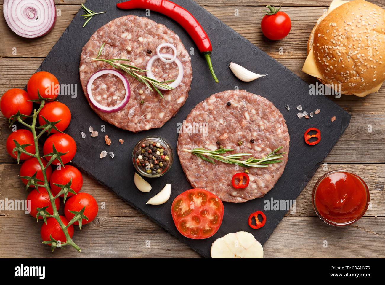 Burgerpasteten mit Rosmarin und Gewürzen auf schwarzem Schieferteller, frische Tomaten, Zwiebeln, Sesambrötchen, Käse. Stockfoto