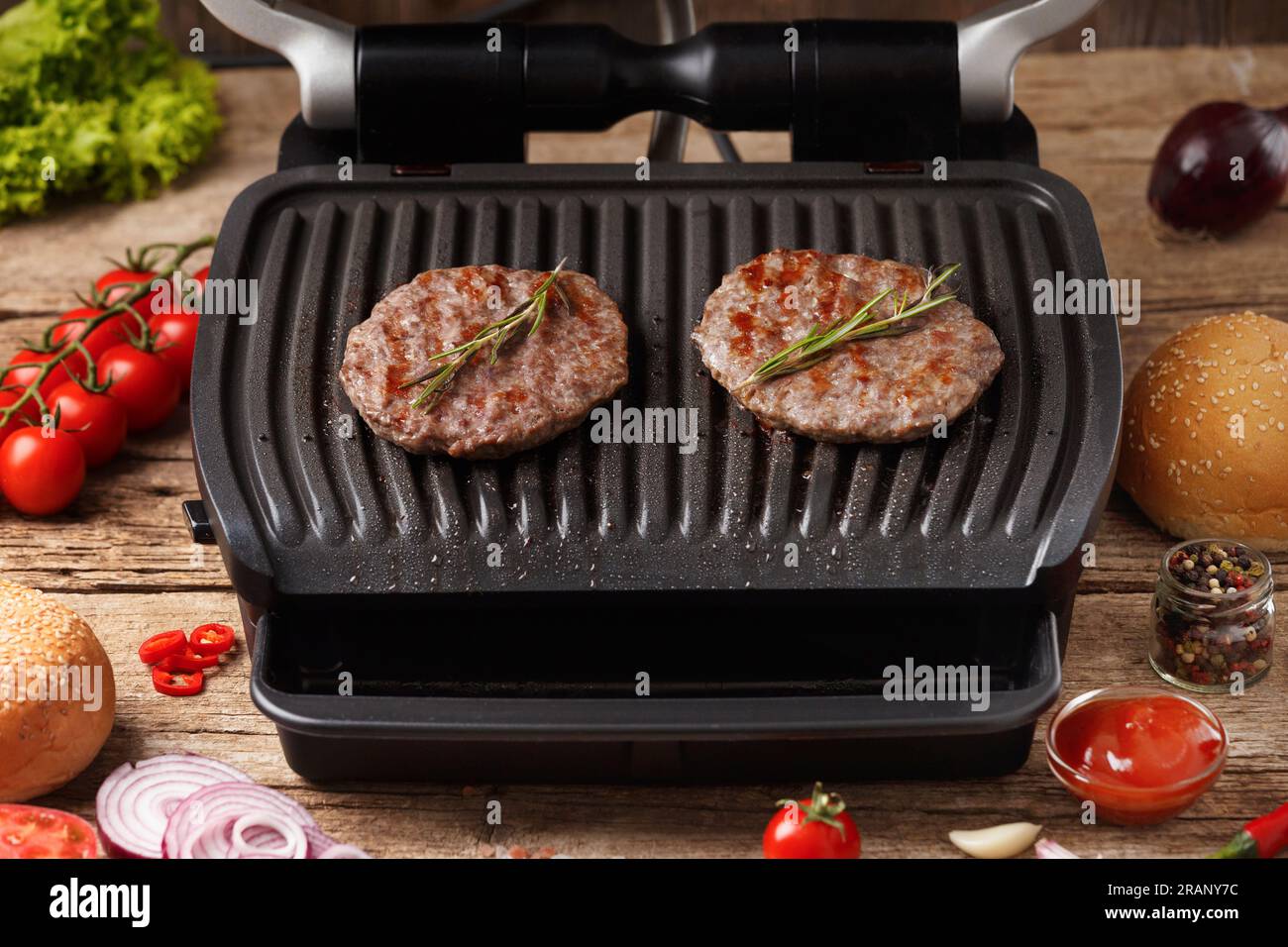 Rinderburger mit Gewürzen werden auf einem elektrischen Grill auf rustikalem Holzhintergrund gebraten. Hausgemachte Hamburger. Zutaten für Burger. Stockfoto