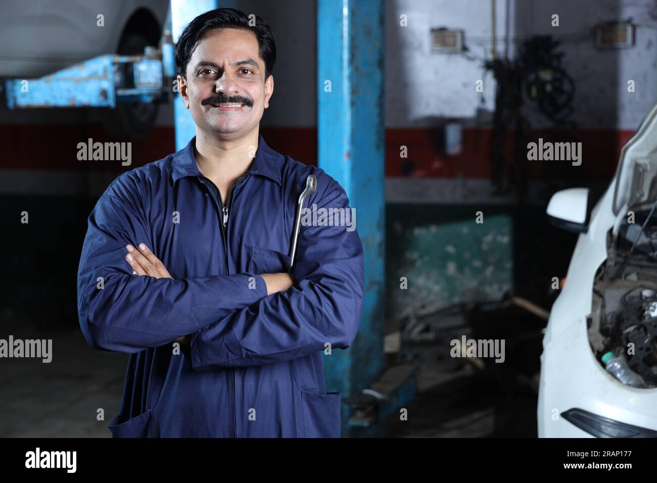 Bärtiger indischer Automechaniker, der beim Autoinspektion lächelt. Ein Autospezialist hält Reparaturwerkzeuge fest. Fachkundiger Mechaniker in Mechanikuniform. Stockfoto