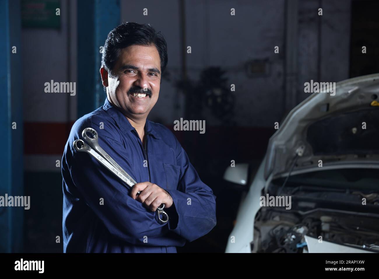 Bärtiger indischer Automechaniker, der beim Autoinspektion lächelt. Ein Autospezialist hält Reparaturwerkzeuge fest. Fachkundiger Mechaniker in Mechanikuniform. Stockfoto