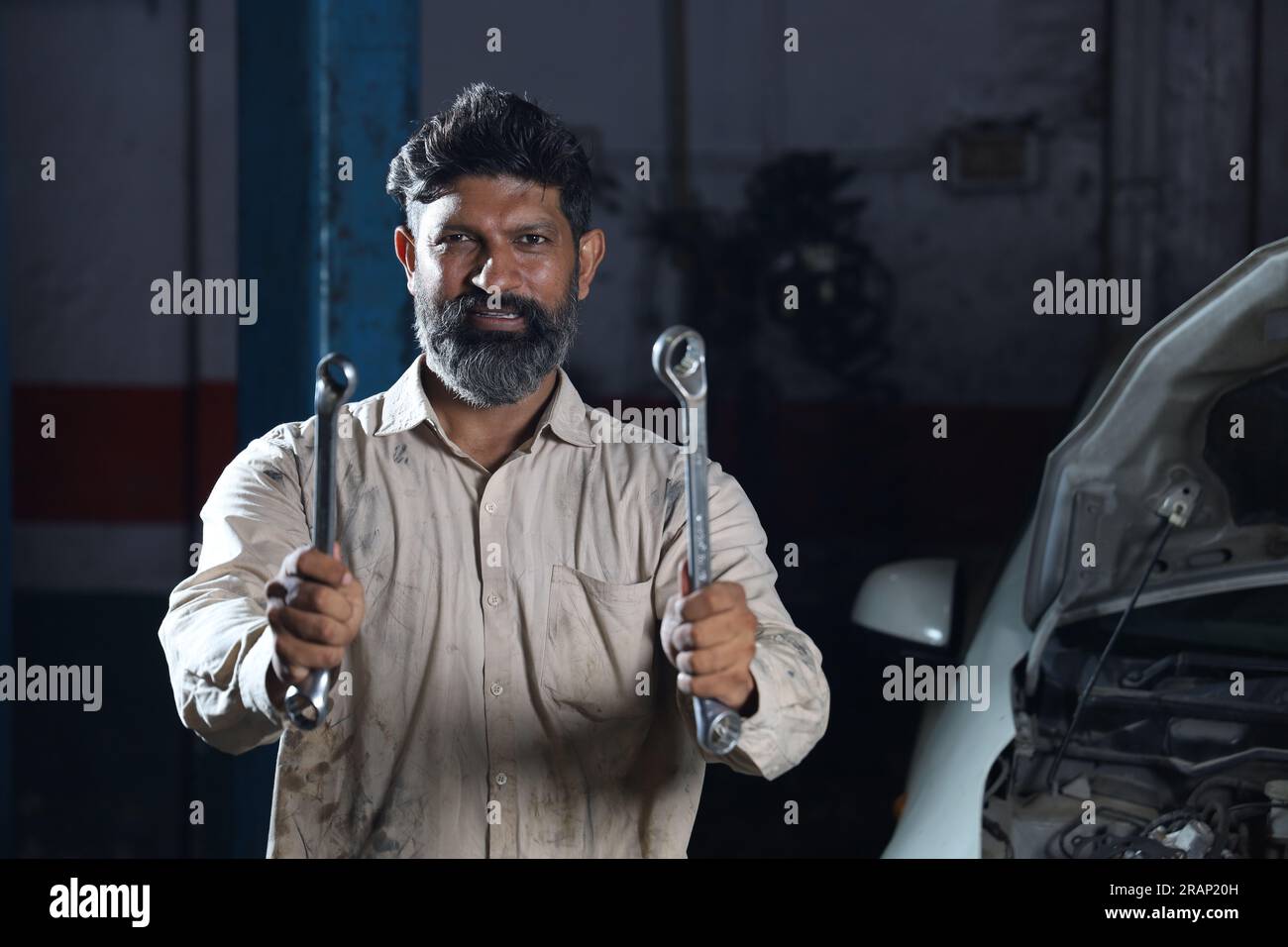 Bärtiger indischer Automechaniker, der beim Autoinspektion lächelt. Ein Autospezialist hält Reparaturwerkzeuge fest. Fachkundiger Mechaniker in Mechanikuniform. Stockfoto