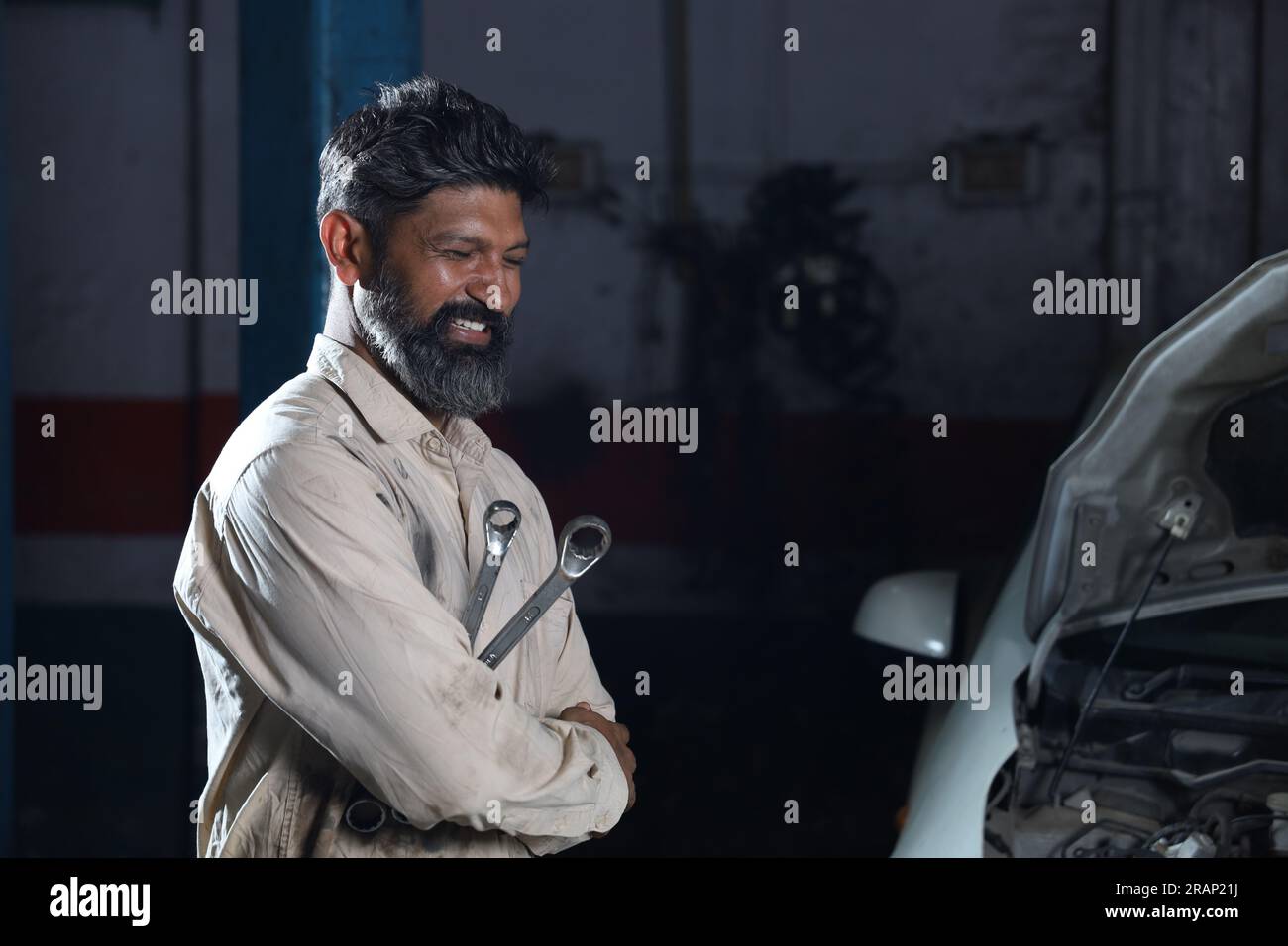 Bärtiger indischer Automechaniker, der beim Autoinspektion lächelt. Ein Autospezialist hält Reparaturwerkzeuge fest. Fachkundiger Mechaniker in Mechanikuniform. Stockfoto