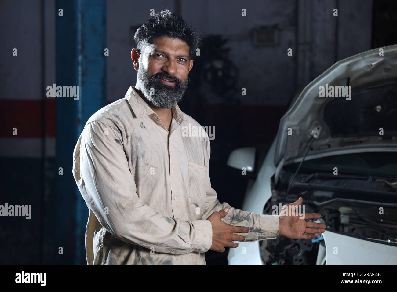 Bärtiger indischer Automechaniker, der beim Autoinspektion lächelt. Ein Autospezialist hält Reparaturwerkzeuge fest. Fachkundiger Mechaniker in Mechanikuniform. Stockfoto