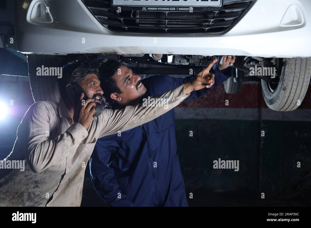Kfz-Mechaniker stehen und arbeiten in einer Tankstelle zusammen. Autospezialisten untersuchen das angehobene Auto. Professionelle Mechaniker telefonieren. Stockfoto