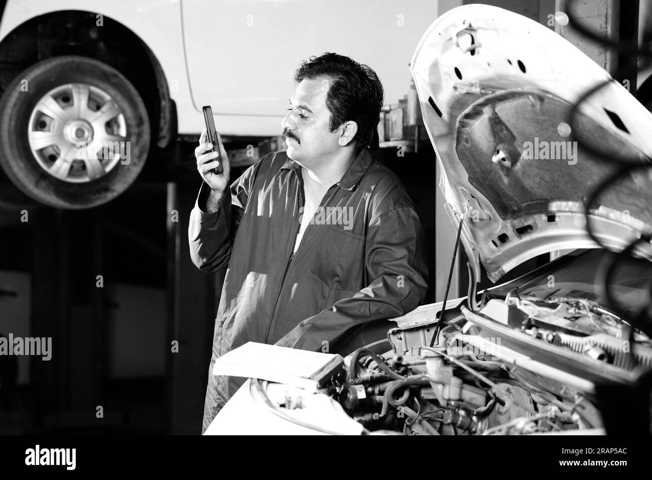 Ein denkender Automechaniker mit Schnurrbart, der seinen Tagesplaner hält, während er das Auto untersucht. Ein Autospezialist steht neben der offenen Motorhaube Stockfoto