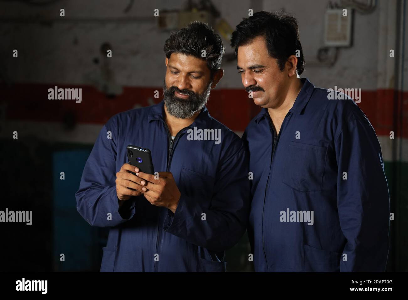 Glücklicher indischer professioneller Automechaniker in Autoreparaturmännern Uniform Surfen auf dem Handy, Smartphone und zusammen lachen, kreative Ideen Stockfoto