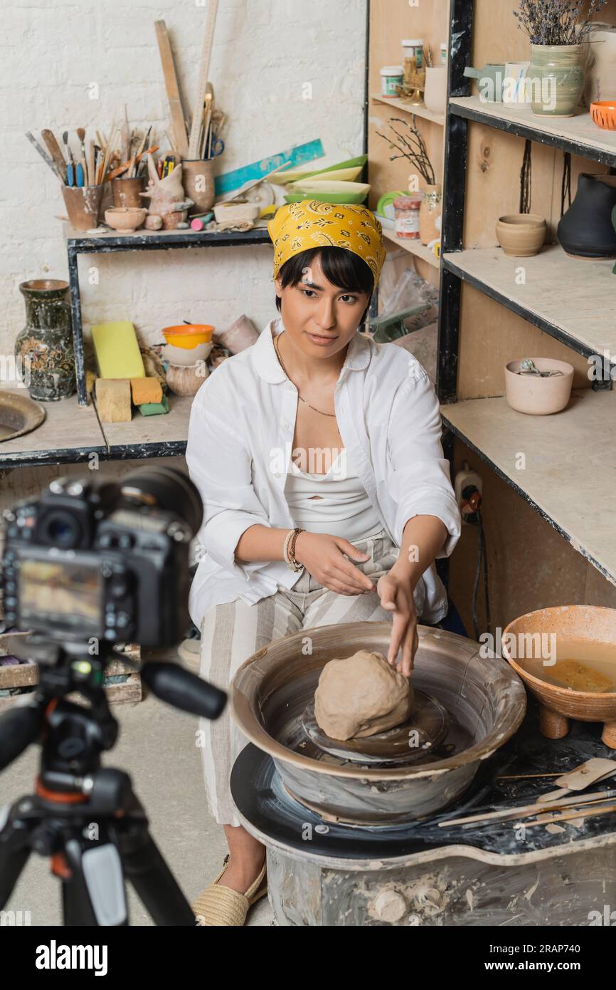 Junge asiatische Künstlerin in Kopftuch und Arbeitskleidung, die auf eine Digitalkamera schaut, während sie in der Keramikwerkstatt am Backgrou neben Ton auf einem Töpferrad sitzt Stockfoto