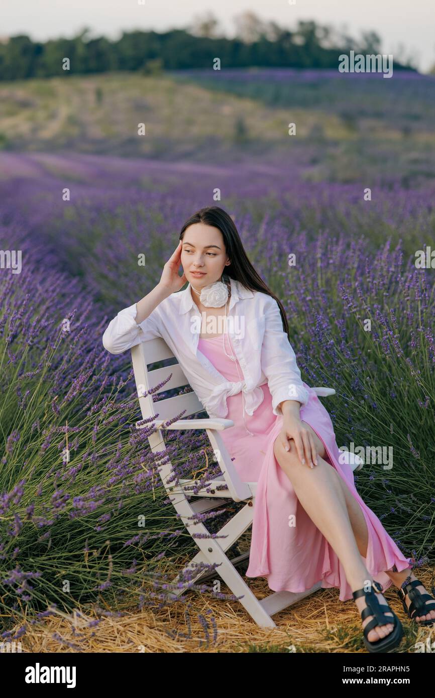 Junge brünette Frau, die in einem Sessel inmitten von blühendem Lavendel sitzt und es vor dem Hintergrund einer wunderschönen Landschaft genießt. Stockfoto