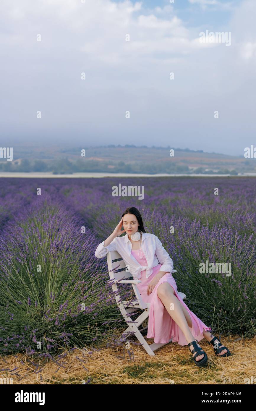 Junge brünette Frau, die in einem Sessel inmitten von blühendem Lavendel sitzt und es vor dem Hintergrund einer wunderschönen Landschaft genießt. Stockfoto