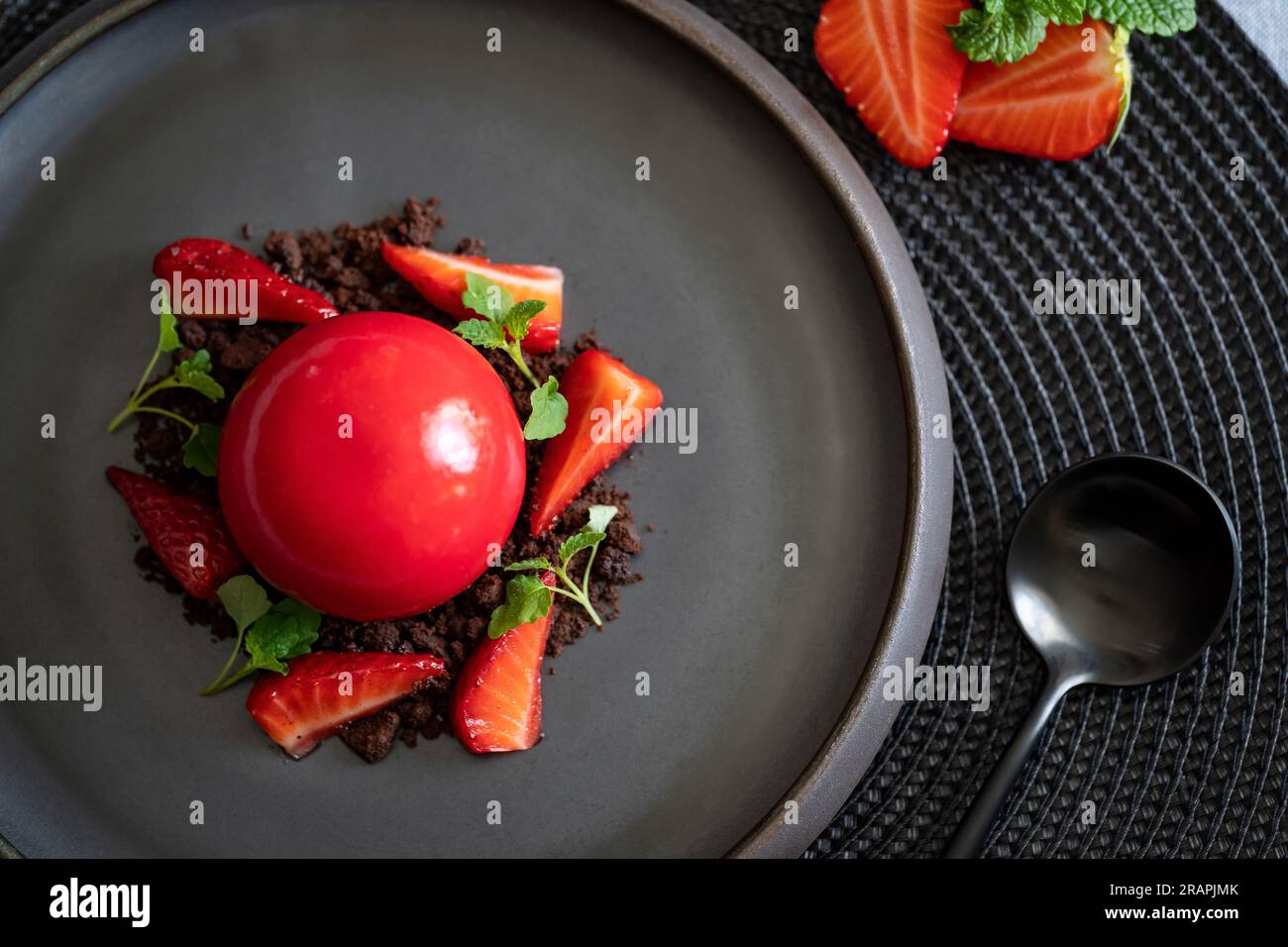 Draufsicht auf einen kugelförmigen Quark mit Erdbeeren und Brownies. Rotes Dessert mit glatten Oberflächen und Spiegelglasur auf dem schwarzen Teller mit schwarzem Löffel. Stockfoto