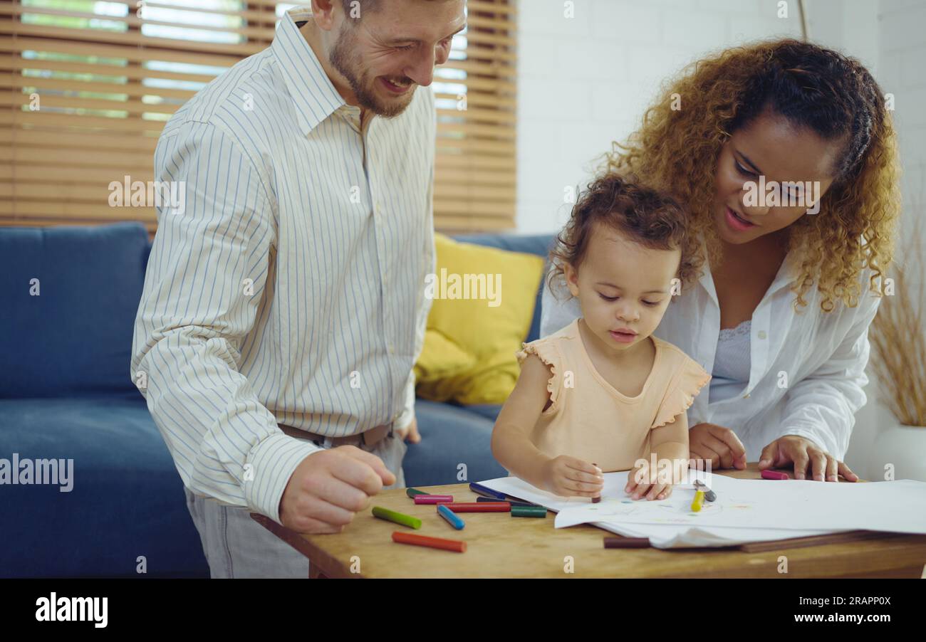 Papa, Mutter und kleine Tochter malen mit bunten Bleistiften auf dem Papier und lächeln fröhlich. Die junge Familie verbringt ihre Freizeit zusammen im Wohnzimmer zu Hause. Stockfoto