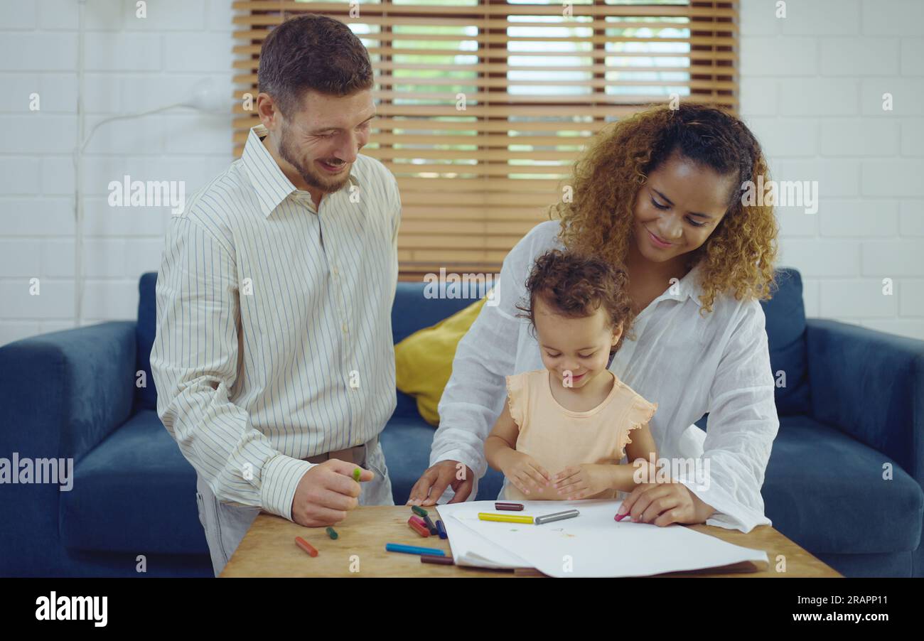 Papa, Mutter und kleine Tochter malen mit bunten Bleistiften auf dem Papier und lächeln fröhlich. Die junge Familie verbringt ihre Freizeit zusammen im Wohnzimmer zu Hause. Stockfoto