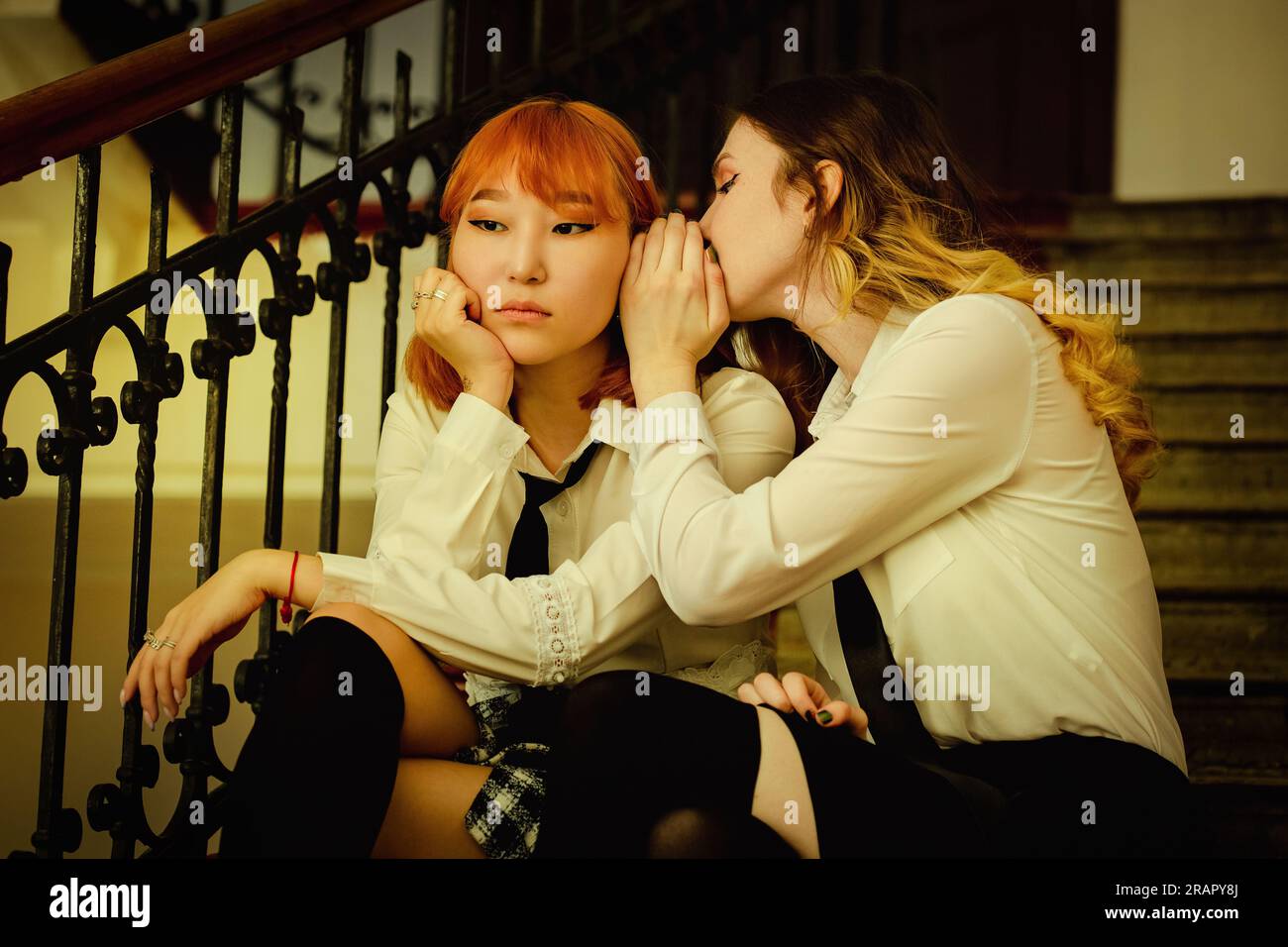 Zwei Schulmädchen in Uniform sitzen auf der Schultreppe. Die süße Blondine flüstert ihr Geheimnis mit ihrer Freundin, einer rothaarigen Asiatin. Freundschaft Stockfoto