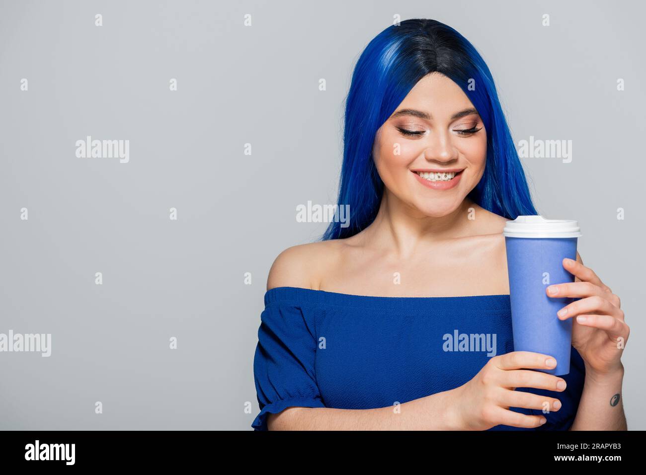 Pappbecher, glückliche junge Frau mit blauen Haaren und Augen, die Kaffee auf grauem Hintergrund hält, Essen zum Mitnehmen, Koffein, Energie, Tattoo, Lebendige Farben Stockfoto