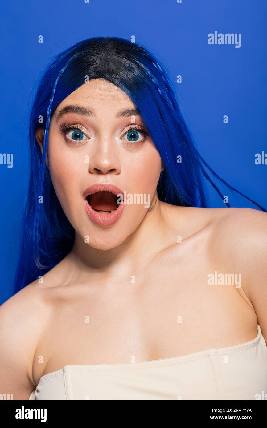 Schockiert, Porträt einer emotionalen jungen Frau mit gefärbtem Haar, Posen auf blauem Hintergrund, Haarfarbe, Individualismus, weibliches Modell mit Make-up und trendy ha Stockfoto