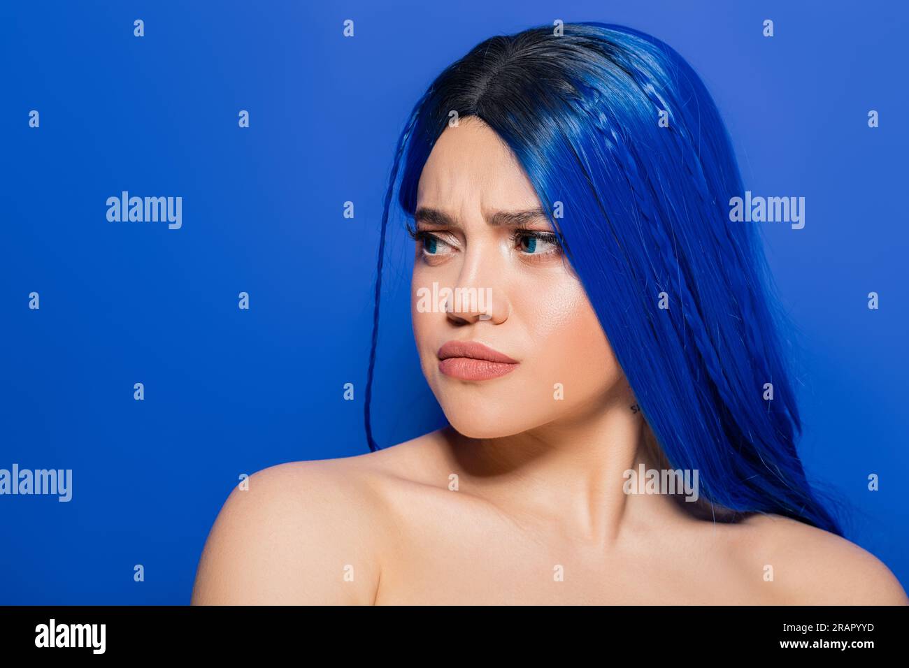 Porträt einer unzufriedenen Frau mit gefärbtem Haar, die auf blauem Hintergrund wegblickt, Haarfarbe, Individualismus, weibliches Modell mit Make-up und trendiges hairstyl Stockfoto