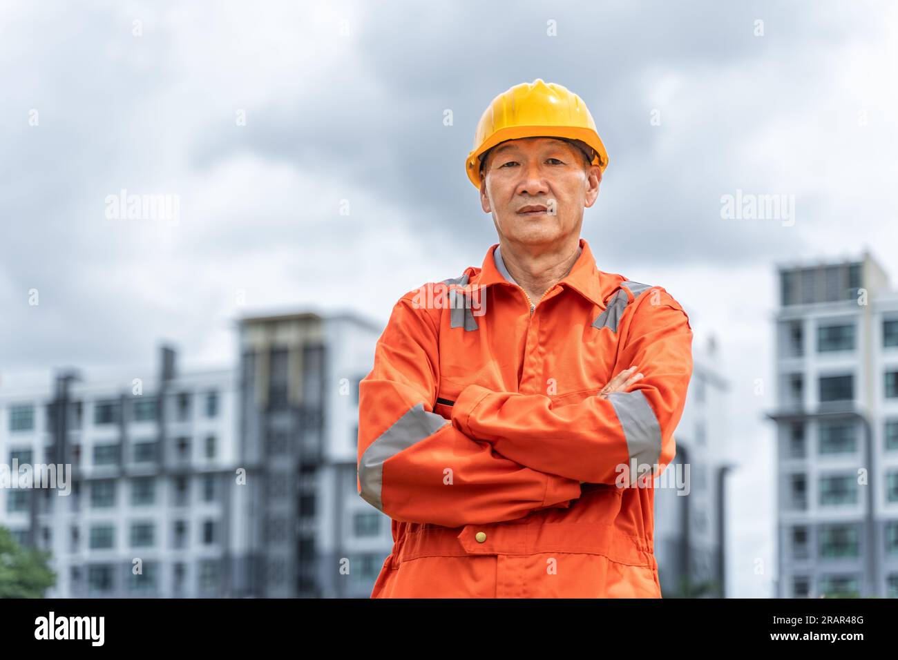 Seriöser und entschlossener asiatischer Ingenieur mittleren Alters in orangefarbenem Overall mit Schutzhelm, der seine Arme kreuzte, während er stolz vor seinem Multi-Stori stand Stockfoto