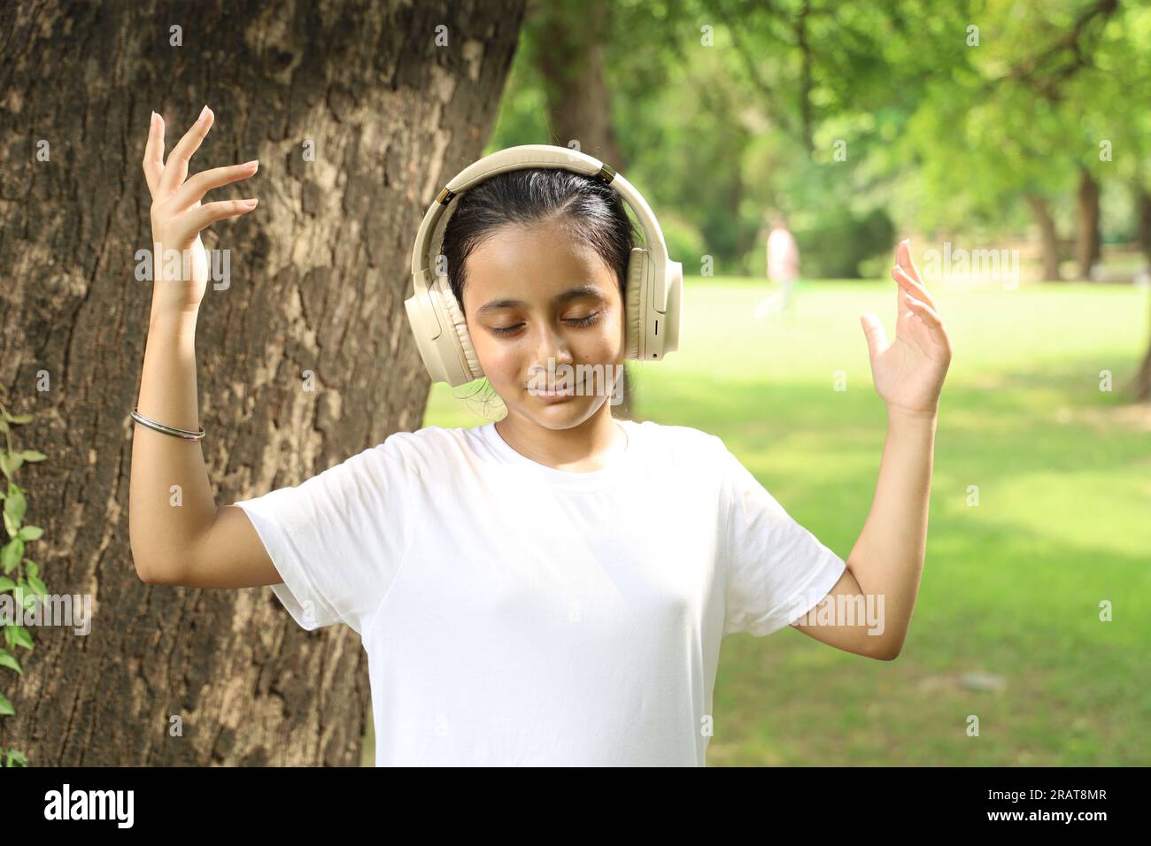 Ein junges Teenager-Mädchen hört und genießt Musik in einem Park mit ihren Kopfhörern unter dem Baum unter Grün. Melodische Musik in friedlicher Atmosphäre. Stockfoto