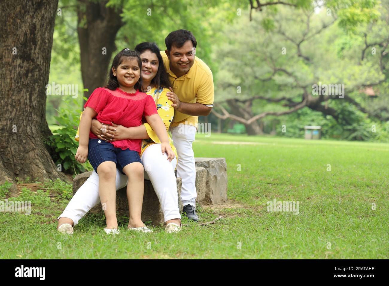 Glückliche indische Familie mit einem Mädchen, das zusammen auf dem Gras sitzt und ihre Familienzeit am frühen Morgen genießt. Die Familie schaut in die Kamera. Stockfoto