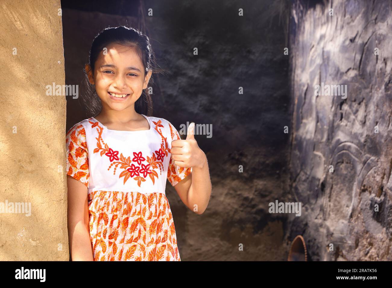 Ein glückliches indisches Mädchen, das im Sommer tagsüber in die Kamera schaut und ein Kleid trägt, auf dem Landhaus steht und lächelt. Daumen hoch mit Lächeln. Stockfoto