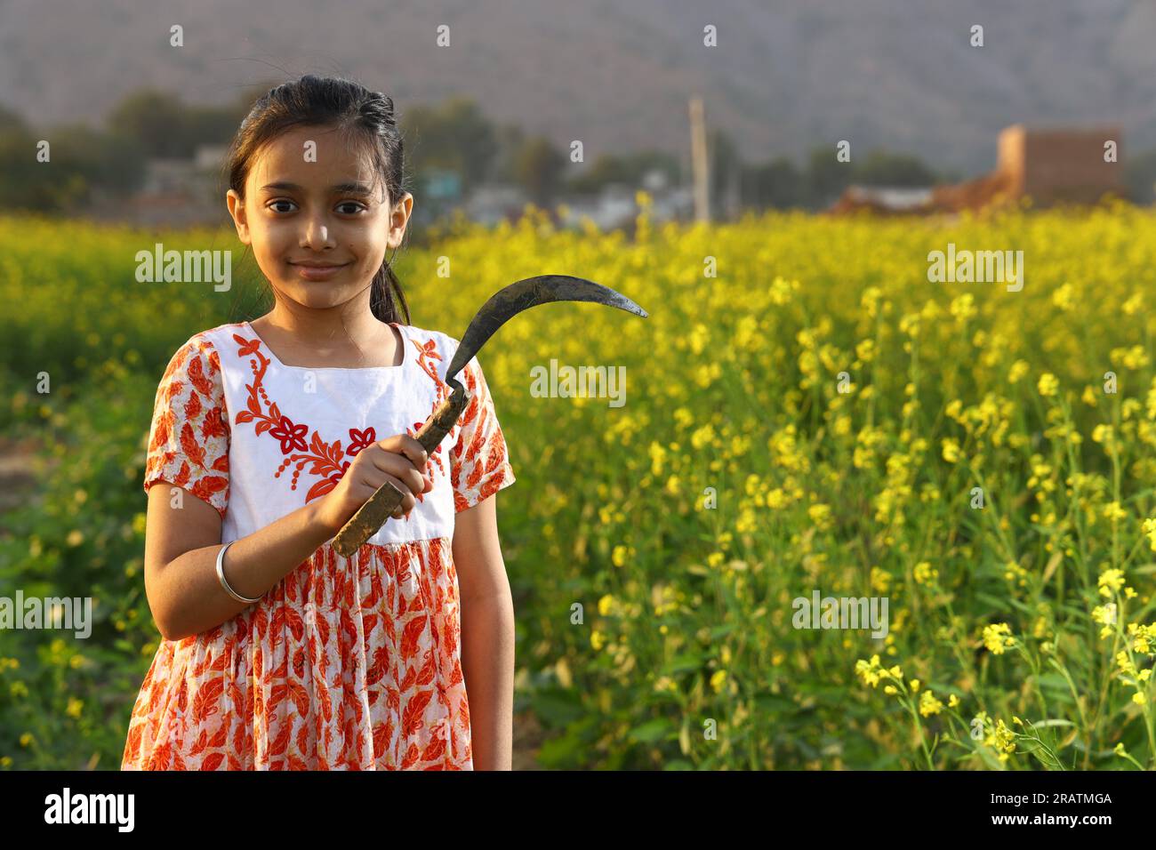 Ein indisches Bauernmädchen, das auf dem Senffeld steht und ein Kleid trägt. Ein Mädchen genießt die blühende Ernte mit Sichel in der Hand. Stockfoto