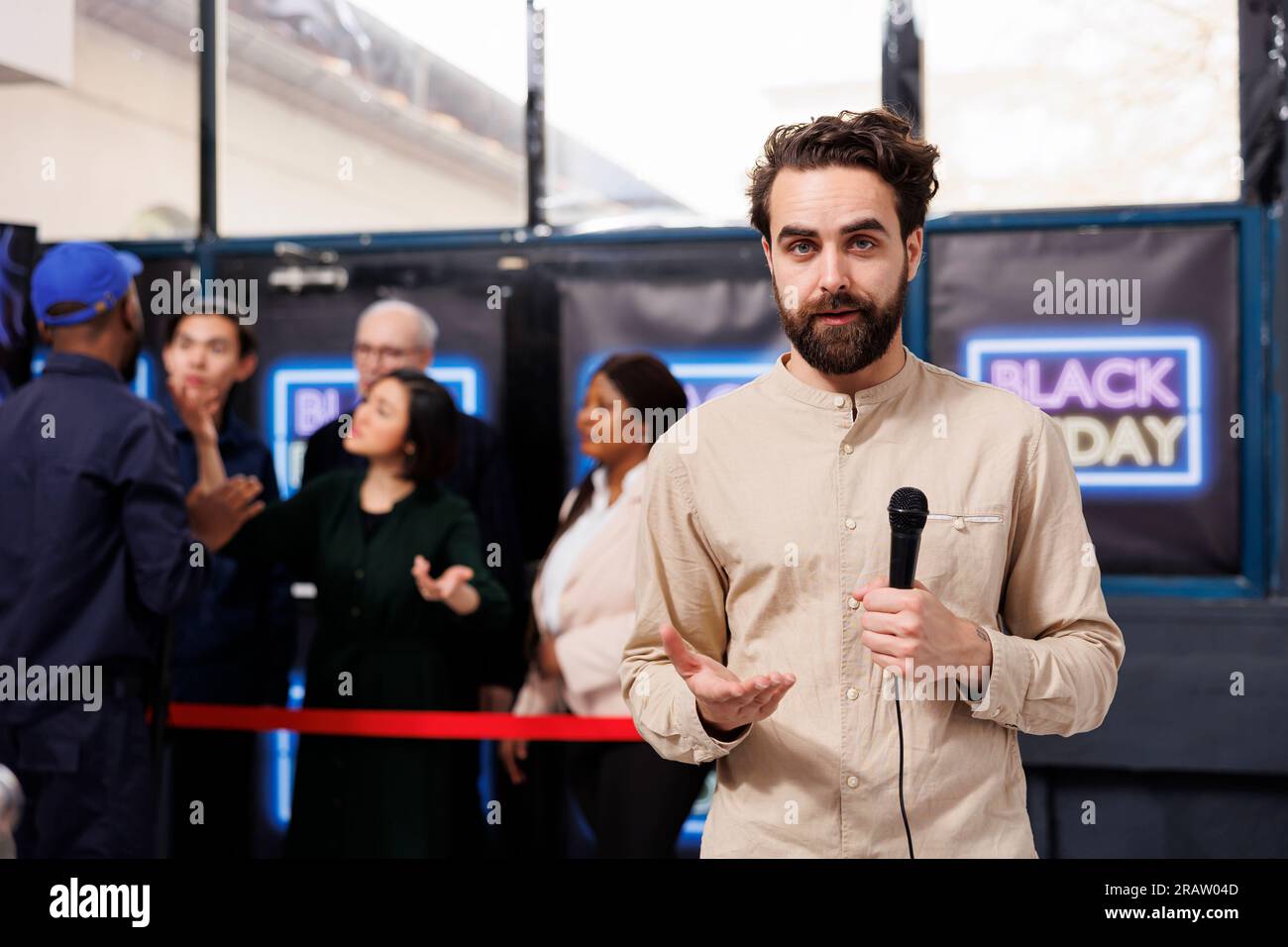 Männlicher TV-Reporter, der während des Black Friday-Verkaufs im Einkaufszentrum steht und Nachrichten für das Fernsehen macht. Ein junger Journalist hält ein Mikrofon in der Hand und spricht mit der Kamera, während er sich gegen die Menge von Käufern stellt Stockfoto