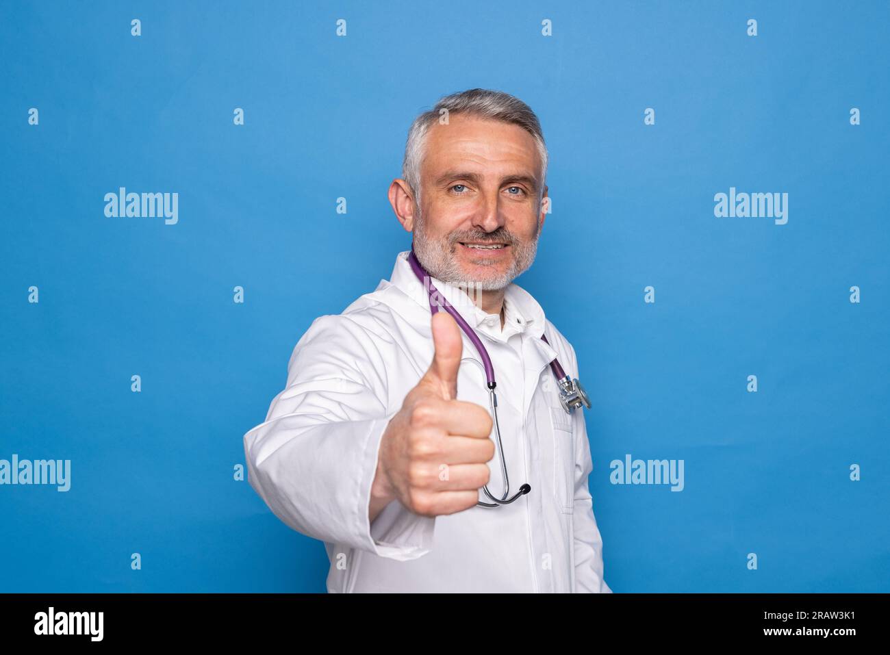 Gut aussehender, fröhlicher, gut aussehender, gut aussehender Mann mittleren Alters in medizinischer Arbeitskleidung Arzt zeigt Daumen hoch und lächelt vor der Kamera, isoliert auf blauem Rücken Stockfoto