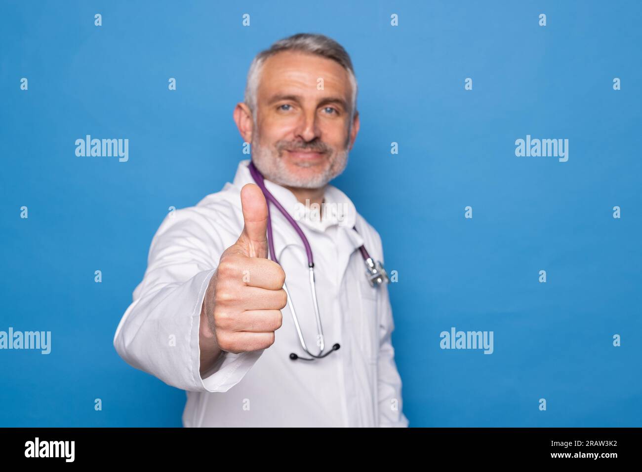 Gut aussehender, fröhlicher, gut aussehender, gut aussehender Mann mittleren Alters in medizinischer Arbeitskleidung Arzt zeigt Daumen hoch und lächelt vor der Kamera, isoliert auf blauem Rücken Stockfoto
