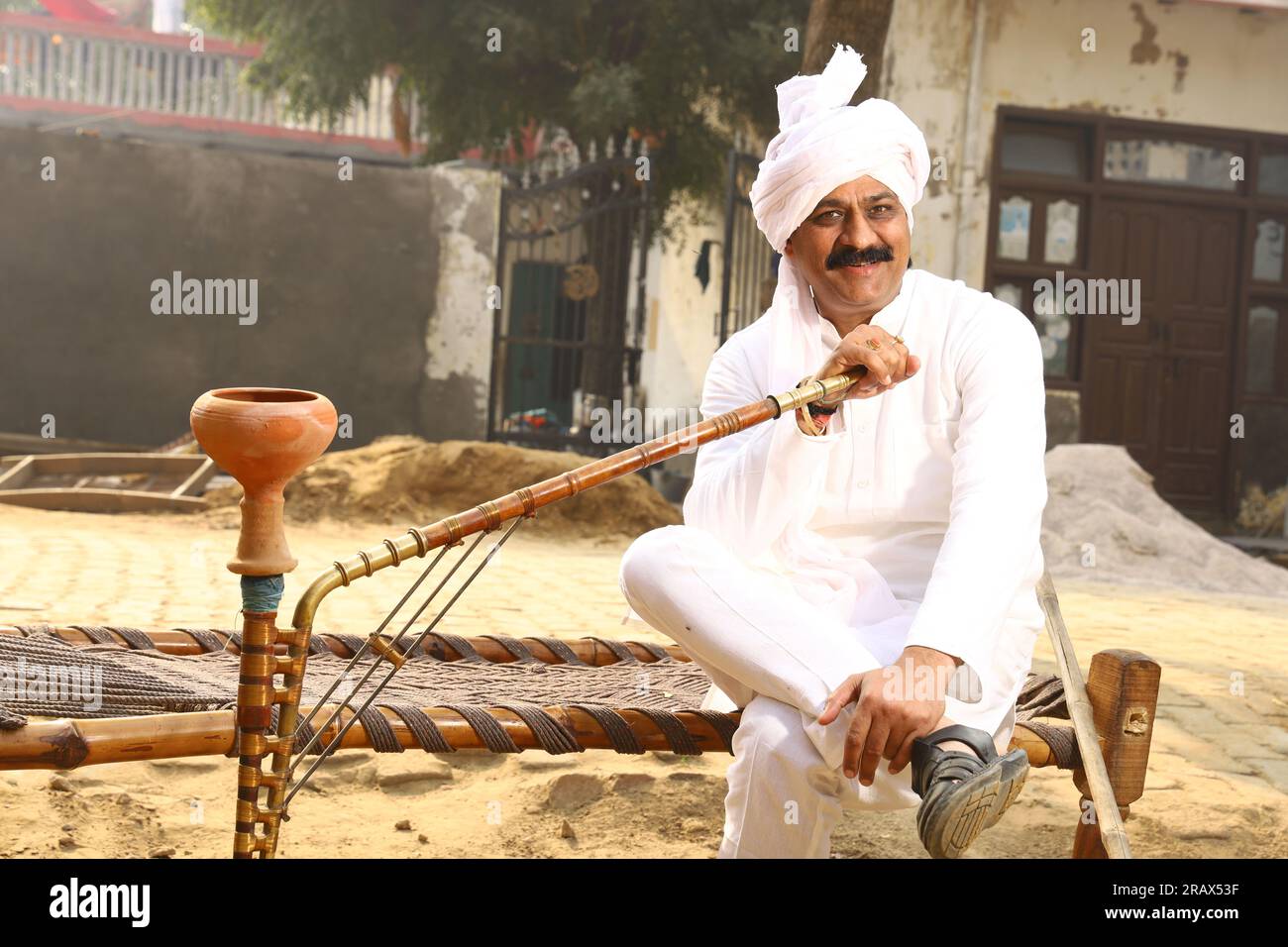 Ein glücklicher Mann aus dem ländlichen Indianerdorf im Turban, der tagsüber draußen sitzt. Hookah in der Hand. Stockfoto