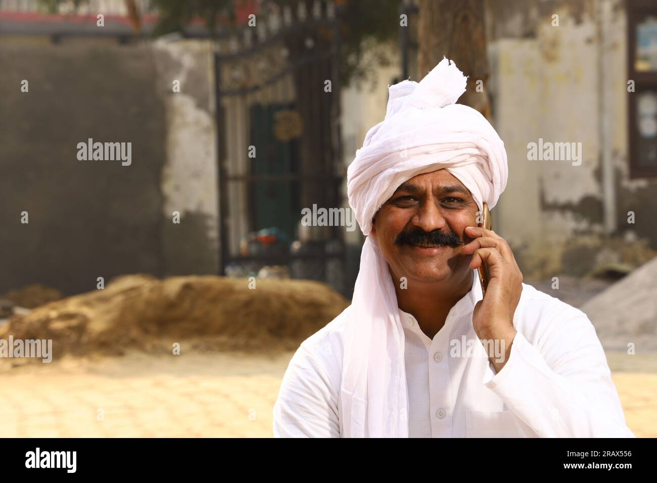 Ein glücklicher Mann aus dem ländlichen Indianerdorf im Turban, der tagsüber draußen sitzt. Telefonieren mit dem Handy. Stockfoto