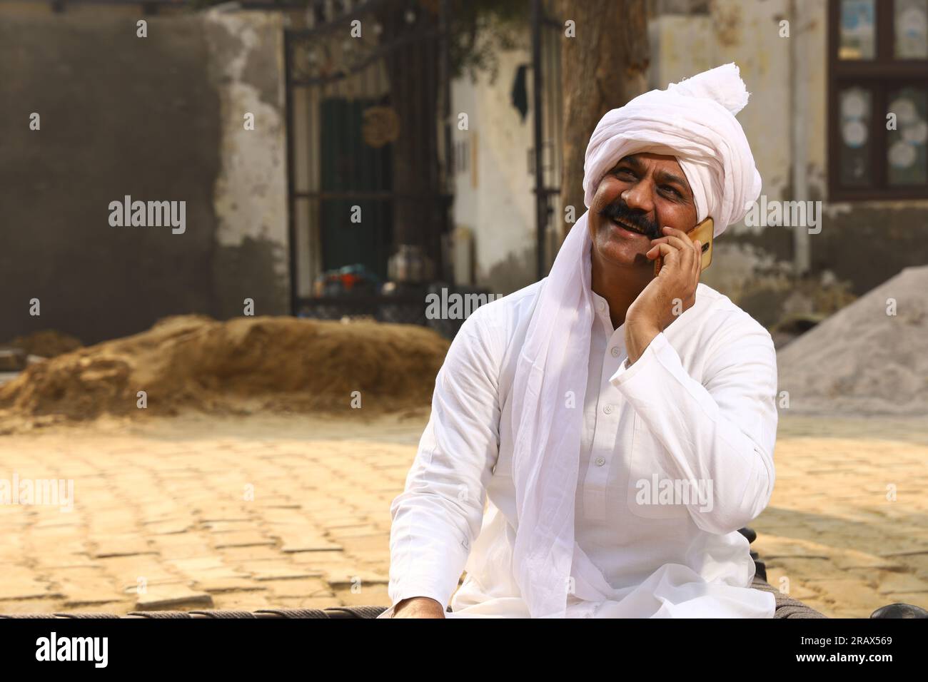 Ein glücklicher Mann aus dem ländlichen Indianerdorf im Turban, der tagsüber draußen sitzt. Telefonieren mit dem Handy. Stockfoto