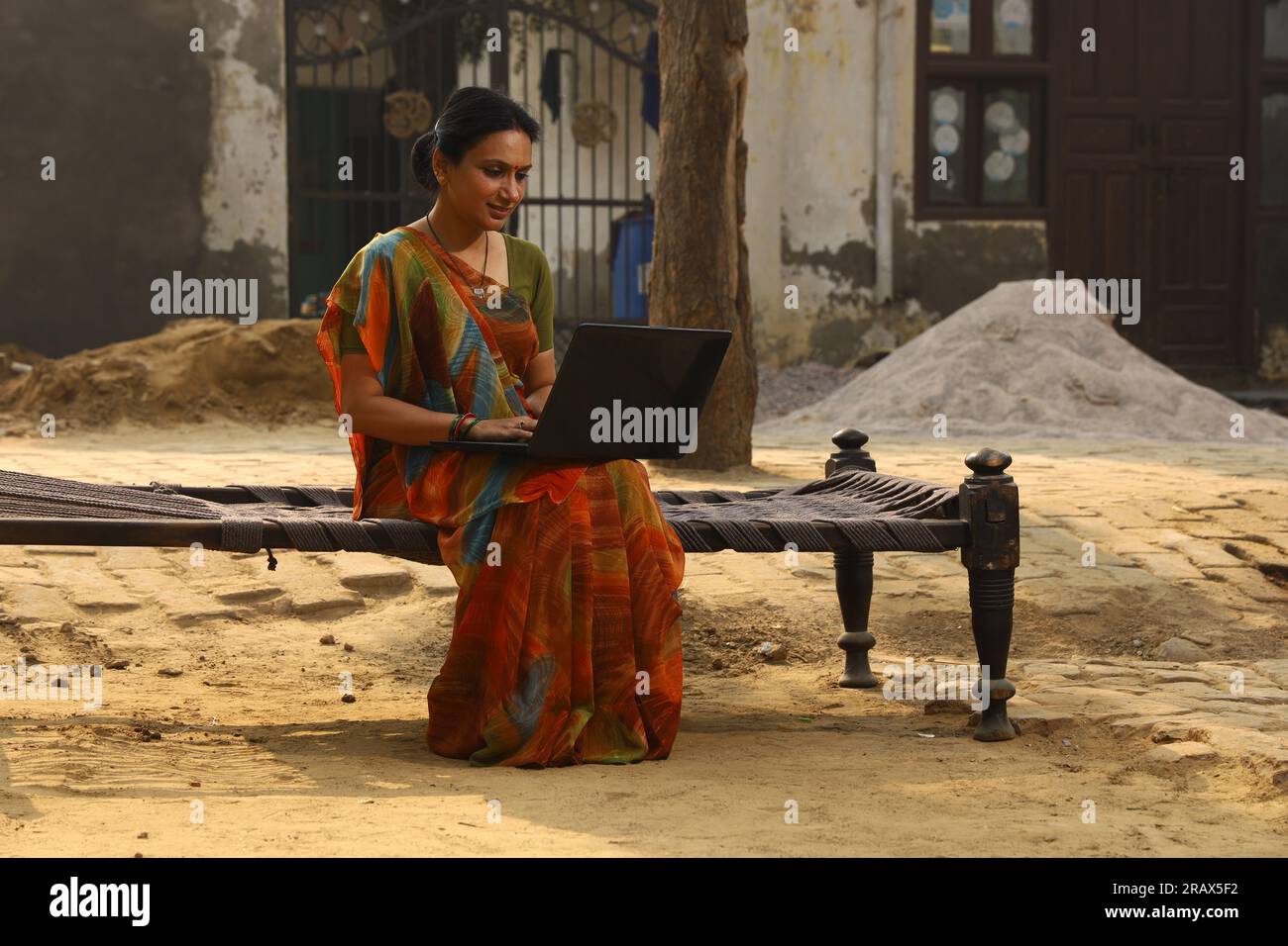 Glückliche indische Landfrau, die auf einem Kinderbett vor ihrem Haus im Vorgarten sitzt. In einem wunderschönen Saree. Arbeiten an einem Laptop. Stockfoto