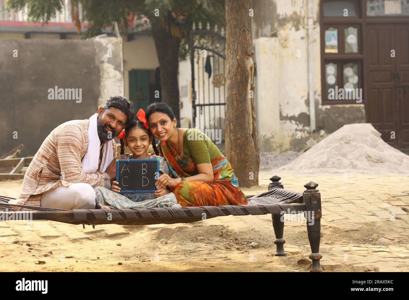 Glückliche ländliche indische Familie, die tagsüber vor ihrer Hütte zusammen saß und ein kleines Whiteboard hielt. Mädchen-Kind-Erziehungskonzept. Stockfoto