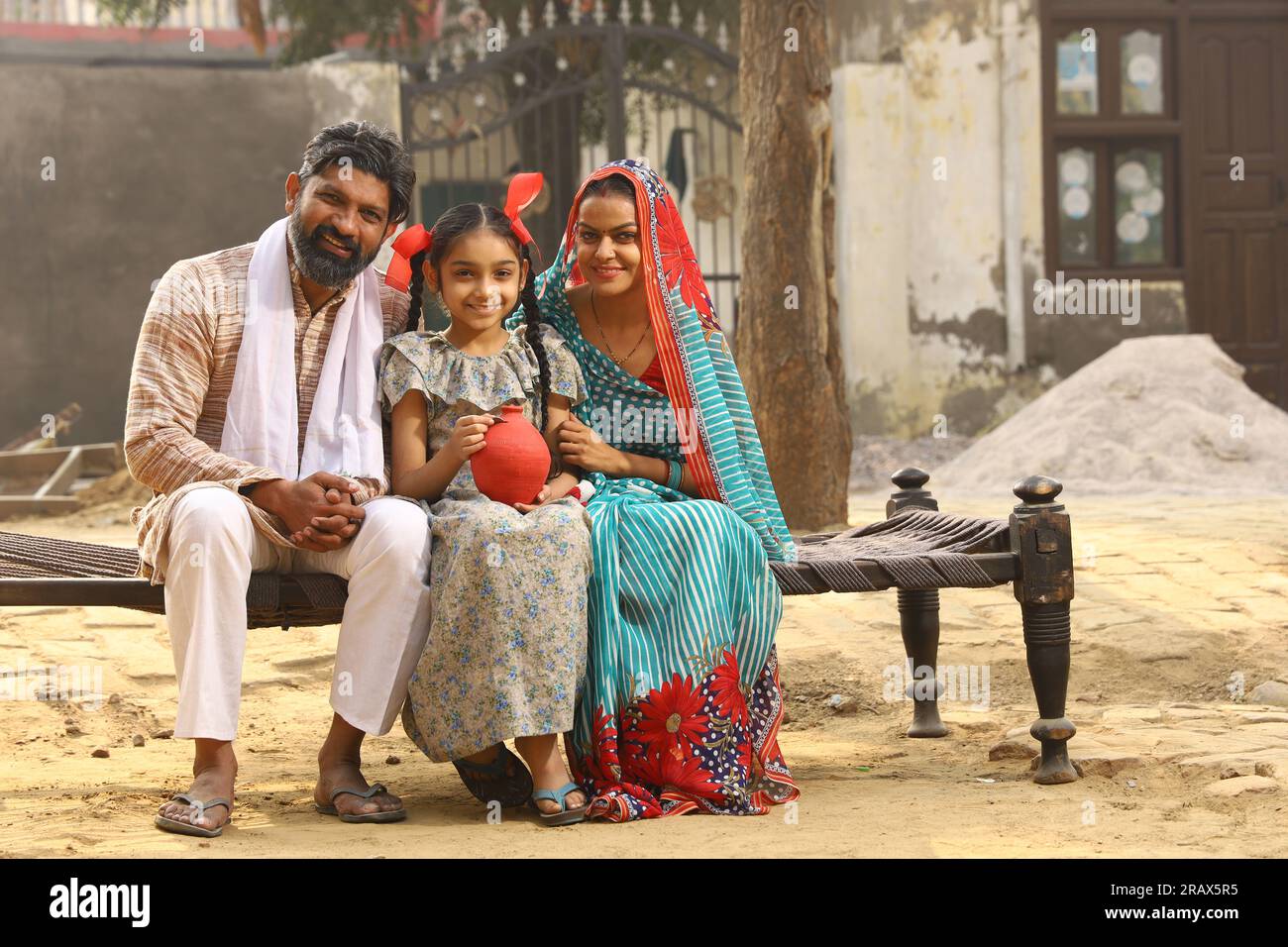 Glückliche ländliche indische Familie, die zusammensitzt, hält die Tochter ein Sparschwein in der Hand, steckt eine Münze hinein und zeigt das Konzept der Geldeinsparung Stockfoto