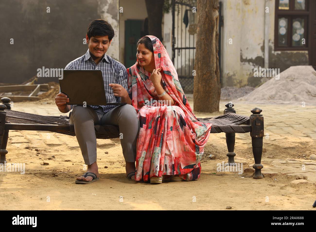 Glückliche indische Landsmutter und Sohn sitzen auf einem Kinderbett im Vorgarten. Mutter in wunderschöner Saree. Arbeiten an einem Laptop. Stockfoto